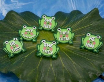 Pode incluir: Seis broches de sapo verde com diferentes opções de pronomes. Os broches estão em uma folha verde. Os pronomes nos broches são: "Ela Ela", "Quaisquer pronomes", "Eles Elas", "Ele Ele", "Ele Eles", e "Ela Elas".