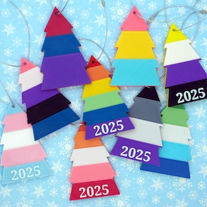 May include: A collection of colorful, layered Christmas tree ornaments. Each tree is a different color combination, with a silver string for hanging. The year "2025" is printed on the bottom of each tree.