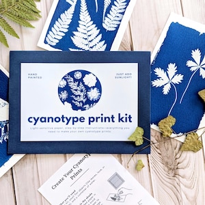 May include: A cyanotype print kit with a navy blue box and several blue and white prints. The kit includes light-sensitive paper and instructions. The prints feature botanical designs and an owl illustration. The text on the box reads "cyanotype print kit".