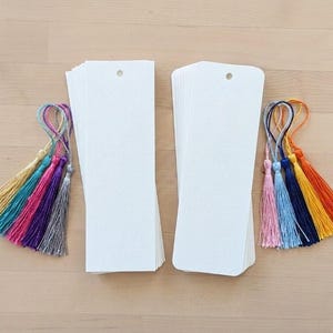Op de afbeelding: Twee stapels rechthoekige bladwijzers van wit karton, elk met een gaatje aan de bovenkant. De ene stapel gaat vergezeld van een set veelkleurige zijden kwastjes in goud, groenblauw, magenta, paars en zilver. De andere stapel bevat roze, lichtblauwe, marineblauwe, gele en oranje kwastjes.