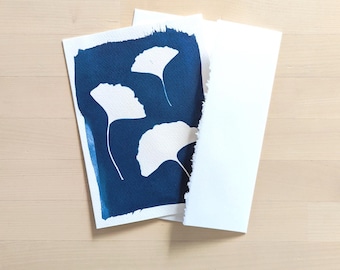 May include: A cyanotype print featuring white ginkgo leaf silhouettes against a deep blue background. The print is on textured paper, with a torn edge, and is accompanied by a blank white card. The artwork is displayed on a light wood surface.