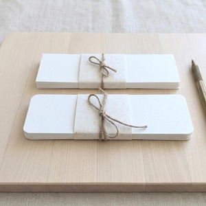 May include: Two stacks of rectangular white cards, wrapped with textured paper and tied with twine bows, sit on a light wooden surface. A brass pen rests nearby. The cards have rounded corners.