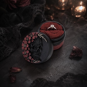 May include: A round black ring box featuring a sculpted skull and red floral relief on the lid. The interior is lined with deep red velvet and holds a silver ring with a single clear stone. The box has a matte finish and intricate detailing.