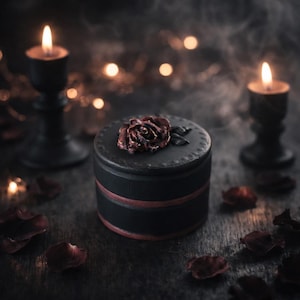 May include: A round black lidded box featuring a dark red rose sculpture on the top surface. The container has a smooth matte finish with a thin horizontal red band encircling the middle section, creating a simple and functional storage vessel.