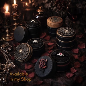 May include: Several round black ring boxes with decorative lids featuring intricate metal filigree, a central gemstone, or a sculpted rose. Each box contains a ring nestled in black fabric. The text Available in my Shop is visible.