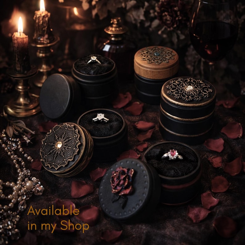 May include: Several round black ring boxes with decorative lids featuring intricate metal filigree, a central gemstone, or a sculpted rose. Each box contains a ring nestled in black fabric. The text Available in my Shop is visible.
