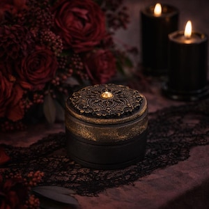 May include: A small, round black box with a decorative, ornate bronze lid. The lid features a central jewel and intricate patterns. The box sits on a dark surface with a lace trim, with lit candles and red flowers in the background.