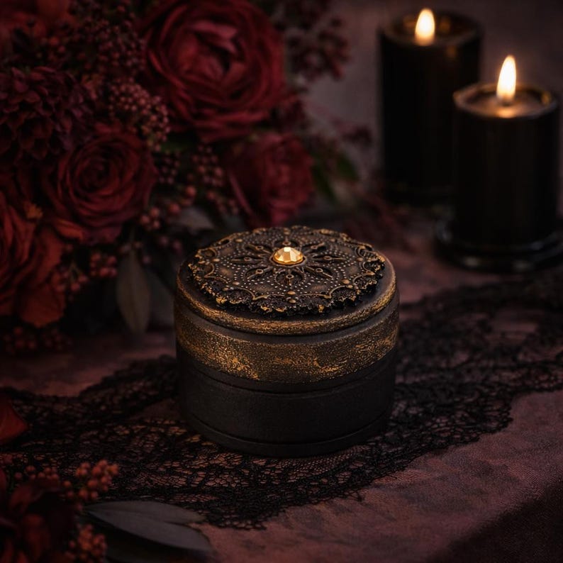 May include: A small, round black box with a decorative, ornate bronze lid. The lid features a central jewel and intricate patterns. The box sits on a dark surface with a lace trim, with lit candles and red flowers in the background.