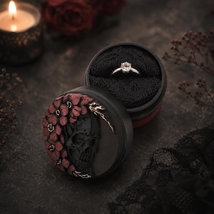 May include: A round black ring box featuring a sculpted black skull relief adorned with dark red flowers and gold accents. The open lid reveals a silver-toned ring with a single clear stone set into a black foam insert inside the container.