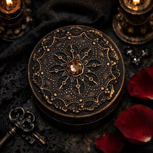 May include: A round, black jewelry box with intricate gold filigree and a central amber-colored gem. The box has a floral design and is surrounded by candles, keys, and rose petals, creating a gothic or vintage aesthetic.