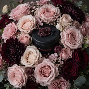 May include: A black cylindrical container with a dark floral accent on the lid, surrounded by a dense arrangement of light pink roses, deep burgundy dahlias, and small filler flowers, creating a rich contrast between the dark center and petals.