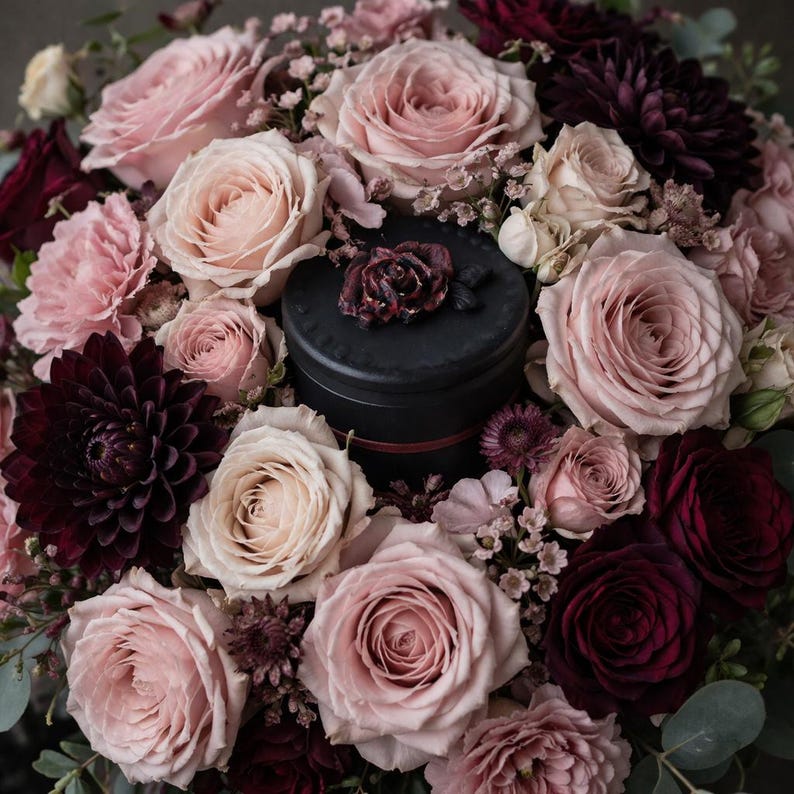 May include: A black cylindrical container with a dark floral accent on the lid, surrounded by a dense arrangement of light pink roses, deep burgundy dahlias, and small filler flowers, creating a rich contrast between the dark center and petals.
