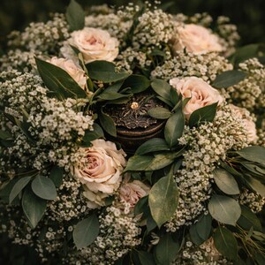 May include: A decorative, antique-style jewelry box with a gold-colored gem on top, nestled within a bouquet of blush-colored roses, baby's breath, and green foliage. The arrangement is captured in a close-up shot, highlighting the textures and colors.