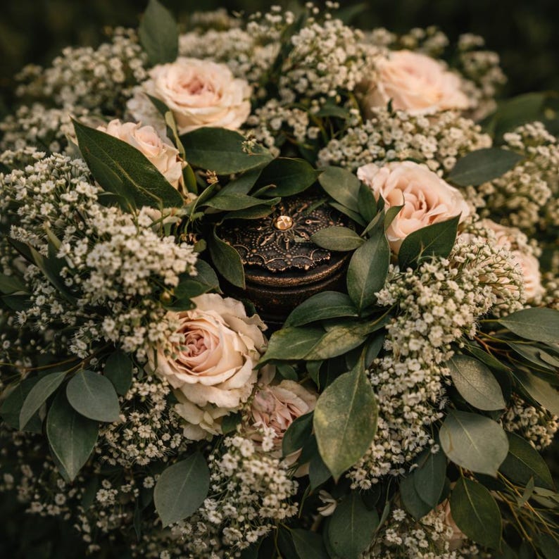 May include: A decorative, antique-style jewelry box with a gold-colored gem on top, nestled within a bouquet of blush-colored roses, baby's breath, and green foliage. The arrangement is captured in a close-up shot, highlighting the textures and colors.