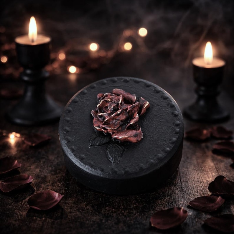 May include: A round black container featuring a three-dimensional metallic copper-colored rose emblem on the lid. The lid has a textured surface with a dotted border, and the overall design is dark and circular, centered in the frame.