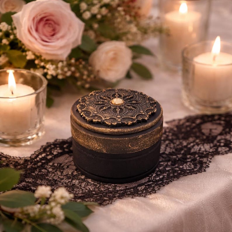 May include: A small, round black box with a decorative, ornate gold lid. The lid features a central jewel and a detailed, floral-like pattern. The box sits on a white surface with black lace and is surrounded by candles and flowers.