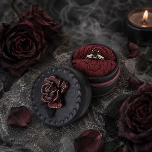 May include: A gold ring with a central black stone and side accents sits inside a round black box with a dark red velvet interior. The lid of the box features a sculpted dark red rose, and the exterior is decorated with a textured border.