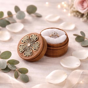 May include: A wooden ring box with a decorative brass flower design and a diamond ring inside. The box is open, revealing the ring nestled in white fabric. The background includes flower petals and greenery.