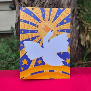 Pode incluir: Um cartão com um cisne branco com as asas abertas contra um fundo de um sol amarelo e raios azuis. O cartão tem uma borda amarela e azul com detalhes de estrelas, sobre uma superfície rosa.