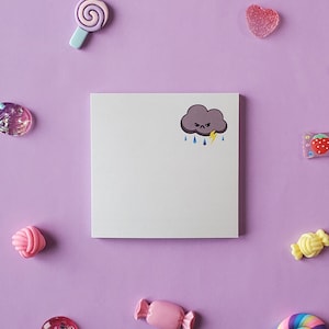May include: A white sticky note with a cartoon illustration of a dark gray cloud with a frowning face, lightning bolt, and raindrops. The note is surrounded by colorful candies and other decorative items on a purple background.
