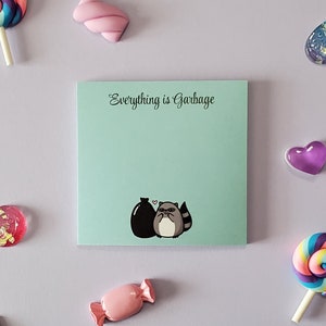 Könnte beinhalten: Ein hellgrüner Haftnotiz mit dem Text "Everything is Garbage" in schwarzer Kursivschrift. Ein Cartoon-Waschbär mit einem Herzen über dem Kopf hält einen schwarzen Müllsack. Der Haftnotiz ist von bunten Süßigkeiten und anderen Dekoartikeln umgeben.