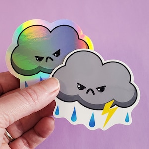 Może przedstawiać: Dwa wzory naklejek z kreskówkową chmurą o złości w oczach. Górna naklejka jest tęczowa, a dolna jest szara z niebieskimi kroplami deszczu i żółtym błyskawicą.