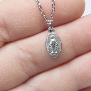 May include: A silver-colored oval pendant necklace featuring a religious figure. The pendant is suspended from a delicate chain. The pendant has an embossed image of a religious figure.