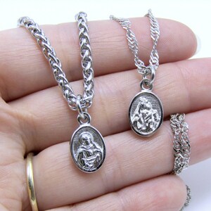 May include: Two silver chains with oval pendants featuring religious figures. The chains are on a hand, with the pendants facing the camera.