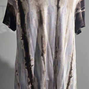 May include: A short-sleeved t-shirt featuring a vertical tie-dye pattern in shades of dark brown, light grey, and white. The garment has a V-neckline and a high-low hemline, with the back portion of the shirt extending longer than the front.