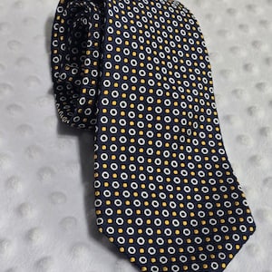 May include: A navy blue necktie featuring a repeating geometric pattern of small white circles and solid yellow dots arranged in a grid. The tie is folded at the top, showing the pointed end and the fabric texture of the woven material.