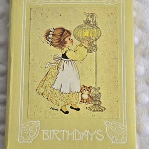 May include: A light yellow hardcover book titled BIRTHDAYS with a white border design. The cover features an illustration of a child in a floral dress and apron standing by a birdcage with a yellow bird, accompanied by a small brown kitten.