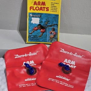May include: A pair of red inflatable arm floats with blue valves and the text Learn-to-Swim Arm Float printed in white. The items are accompanied by a yellow cardboard box featuring the text Learn-to-Swim Arm Floats and Poolmaster branding.