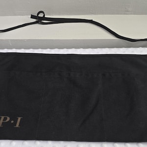 May include: A black waist apron featuring three front pockets and a long tie strap at the top. The text OPI is printed in a dark grey font on the lower left side of the apron. The fabric appears to be a solid black textile material.