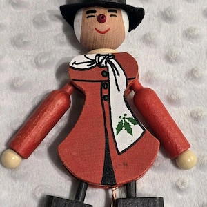 May include: A wooden Christmas ornament shaped like a figure wearing a black top hat with green holly leaves, a red coat with a white scarf featuring green holly, and black shoes. The figure has a round face with a red nose and a painted smile.