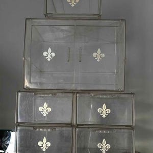 May include: A stack of five clear acrylic storage boxes arranged in a tiered formation. Each box features a white fleur-de-lis emblem centered on the front panel. The boxes are rectangular with transparent walls and integrated handles on the drawers.