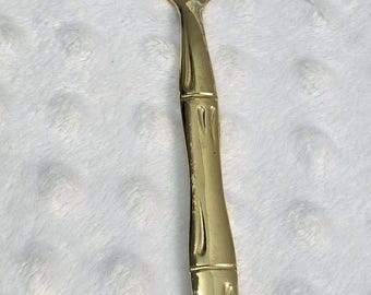 May include: A gold-toned metal cake server featuring a smooth, pointed, oval-shaped blade and a handle designed with a textured, segmented bamboo-like appearance. The utensil has a polished, reflective finish throughout its entire surface.