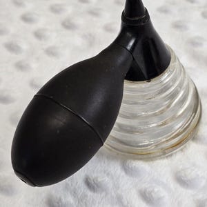 May include: A black rubber squeeze bulb attached to a clear glass base with a ribbed, conical shape. The black nozzle extends upward from the top of the glass section, which features a series of horizontal ridges circling the exterior surface.