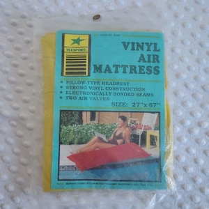 May include: A yellow Texsport vinyl air mattress in clear plastic packaging. The label features the text VINYL AIR MATTRESS, PILLOW-TYPE HEADREST, STRONG VINYL CONSTRUCTION, ELECTRONICALLY BONDED SEAMS, TWO AIR VALVES, and the size 27 inches by 67 inches.