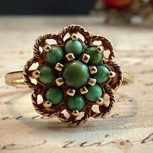 Puede incluir: Un anillo de oro con una piedra verde redonda central rodeada por ocho piedras verdes más pequeñas en forma de flor. El engaste tiene un borde de metal trenzado con detalles calados, creando un diseño circular sobre una banda de oro lisa.