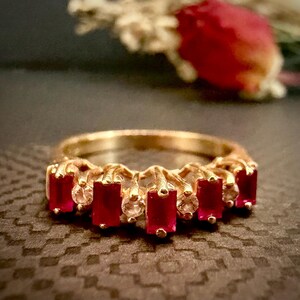 May include: A gold ring featuring a row of alternating rectangular red gemstones and small round clear stones. The band is crafted from a polished gold-toned metal, and the stones are set in a prong setting across the top of the ring.