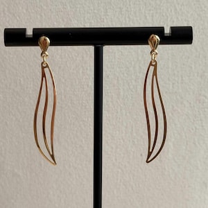 Könnte beinhalten: Ein Paar goldfarbene Ohrhänger mit einem schlanken, länglichen und geschwungenen Design mit offenem Rahmen. Jeder Ohrring besteht aus zwei parallelen, geschwungenen Linien, die sich oben und unten treffen und an einem kleinen, einfachen Haken hängen.