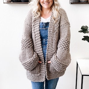 May include: A woman wearing a light brown knitted cardigan with large, puffy sleeves and a denim overall dress. She is standing in front of a white wall with shelves of yarn.