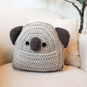 May include: A crocheted koala plush toy with brown ears and a brown nose. The toy has black button eyes and a small pocket on the front. The toy is sitting on a white couch.