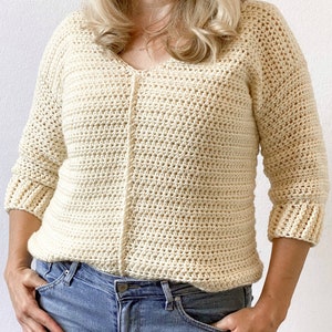 May include: A cream-colored crocheted sweater with a v-neck and three-quarter sleeves. The sweater is worn over blue jeans.