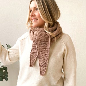 May include: A mauve crocheted scarf with a textured pattern. The scarf is worn around the neck of a person wearing a cream sweater and blue jeans.