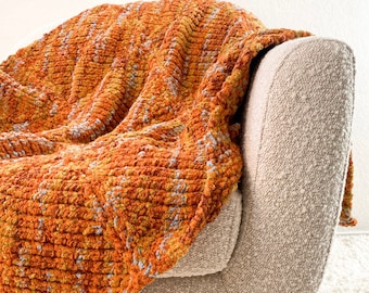 May include: A soft, orange and yellow textured throw blanket draped over a beige armchair. The armchair has a wooden base and is sitting on a white shag rug.
