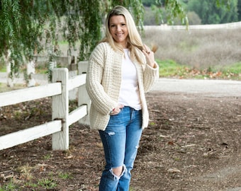 May include: A woman wearing a cream knitted cardigan, a white t-shirt, blue jeans with distressed knees, and black ankle boots. She is standing in front of a white picket fence with a grassy area behind her.