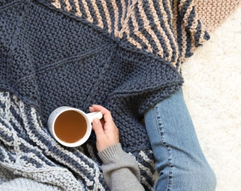 May include: A person is wrapped in a gray, white, and brown striped knitted blanket while holding a white mug of tea. The person is wearing blue jeans and a gray sweater.