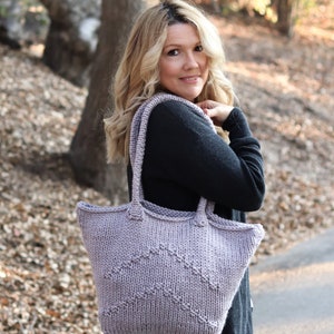 May include: A woman is carrying a large, knitted tote bag with a chevron pattern. The bag is a light purple color and has a wide, comfortable strap.