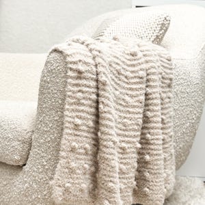 Puede incluir: Una manta beige texturizada sobre el brazo de un sillón crema. La manta tiene un patrón ondulado y rayado con pequeñas protuberancias en relieve. La silla y la manta crean una estética acogedora y de tonos neutros.